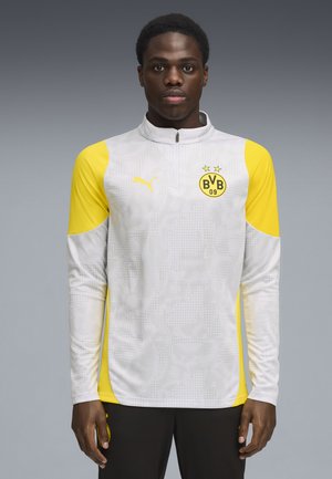 Sudadera deportiva de manga larga en gris claro con acentos amarillos. Presenta un cierre de cuarto de cremallera, tejido texturizado y el logo del equipo en el pecho.