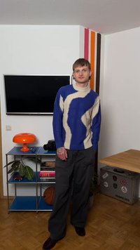 Jeune personne portant un pull à motifs bleu et beige se tient dans une pièce avec une télévision murale, une lampe orange, un ballon de basket et des rayures décoratives sur le mur.