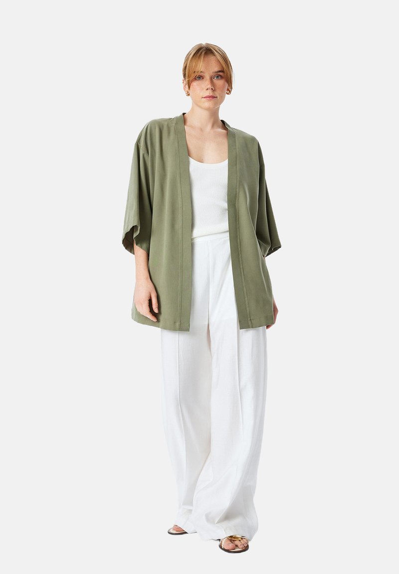 Lichte, olijfgroene kimono-stijl jas met brede mouwen, gecombineerd met een witte tanktop en losse witte broek. Eenvoudige, gestructureerde stof.