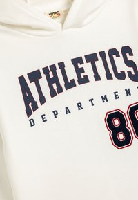 Vit huvtröja med "ATHLETICS DEPARTMENT" i mörkblå och röd text. Har ett stort, röd kontur nummer "80" och en mjuk textur.