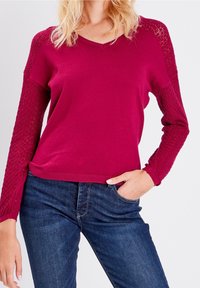 V-ausschnitt Strickpullover in Fuchsia mit strukturierten Spitzenärmeln. Mit gerippten Bündchen und Saum. Kombiniert mit dunkelblauen Jeans.