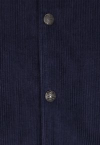 Marineblå corduroy-stof med vertikale ribber, der har tre runde metalknapper med præget tekst til lukning.