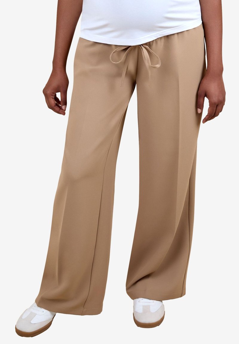 Persona che indossa pantaloni beige a gamba larga con coulisse in vita, sneakers bianchi e top bianco, in piedi davanti a uno sfondo semplice.