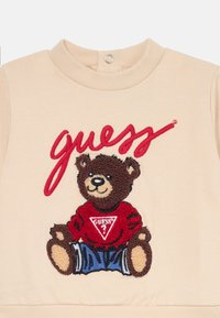 Beige genser med brodert brun bjørn i rød skjorte med trekantet logo. Myk tekstur med rund hals og trykknapp-lukking.