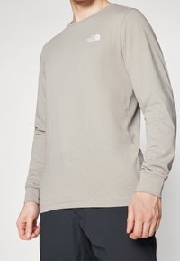 Helles graues Langarmshirt aus weichem Material, mit Rundhalsausschnitt, taillierten Bündchen und einem kleinen weißen Logo auf der Brust.