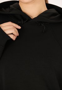 Zwarte hoodie met een trekkoordsluiting, zachte textuur en lange mouwen. De halslijn is rond en de stof lijkt glad en lichtgewicht te zijn.