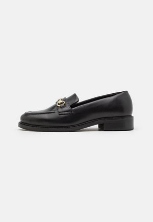Zwarte leren loafer met lage hak en gouden metalen bit-detail op de wreef, zijaanzicht tegen een effen witte achtergrond.