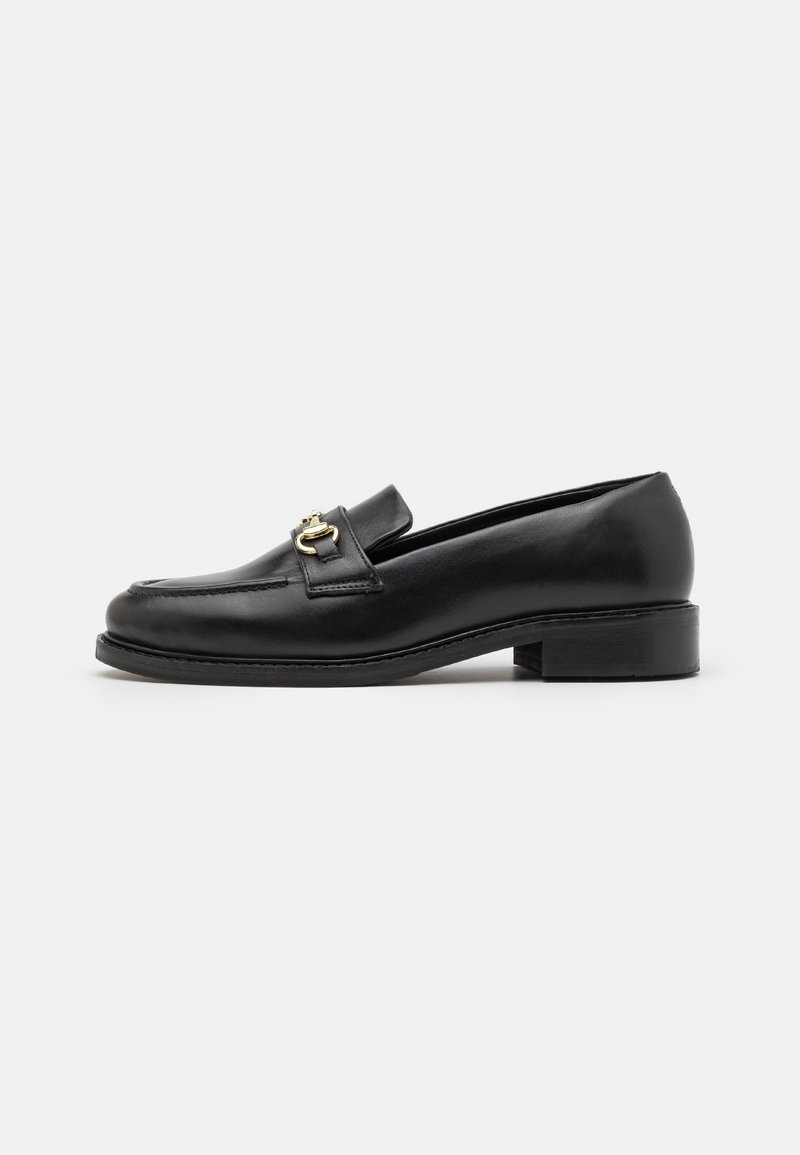 Mocassin en cuir noir avec petit talon et détail en métal doré sur le dessus, présenté de profil sur un fond blanc uni.