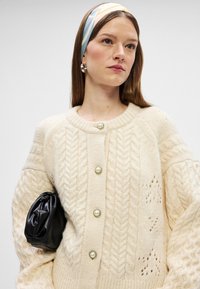 Cárdigan de punto crema con patrones de cable, con botones de aspecto perlado y cuello redondo. Combinado con un clutch negro y una diadema estampada.