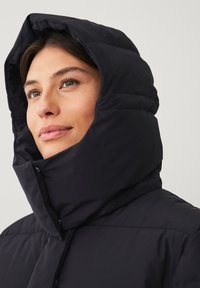Sort pufferjakke med en høj, quiltet krave og hætte. Glat tekstur, synlige syninger og front snap-lukning.