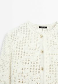 Hvid, strikket cardigan med ternet mønster, rund halsudskæring og knapper i guld-tone foran. Blød tekstur og let design.