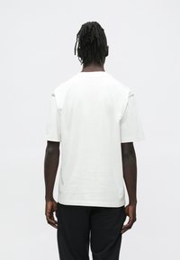 Άνδρας με ντραντλοκ που βλέπει προς τα πίσω, φοράει απλό λευκό κοντομάνικο πουκάμισο και μαύρο παντελόνι μπροστά σε λευκό φόντο.