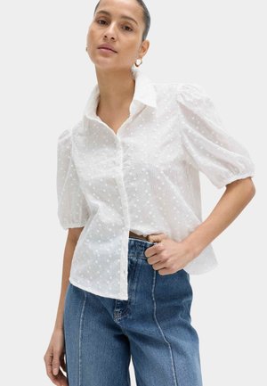 Femme portant un chemisier blanc à manches bouffantes en œillet avec col et un jean bleu taille haute, posant avec une main sur la hanche devant un fond uni.