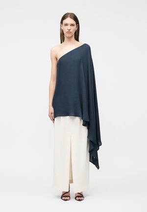 Moteris dėvi tamsiai mėlyną vieno peties laisvai krentančią palaidinę su klostuotu audiniu ir baltas aukšto liemens plačias kelnes su priekiniu plyšiu.