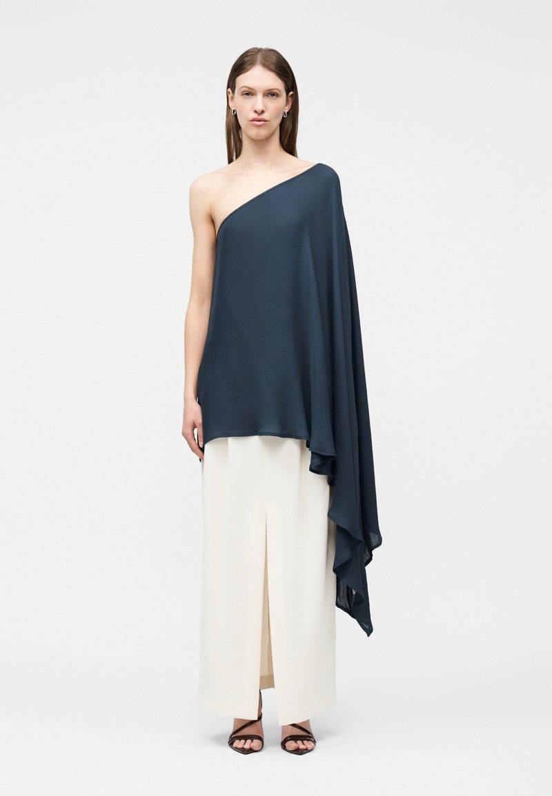 Nő sötétkék, egyszáras, lebernyeges, redőzött anyagú felsőben és fehér, magas derekú, bőszárú nadrágban, amely elöl hasítékkal rendelkezik.