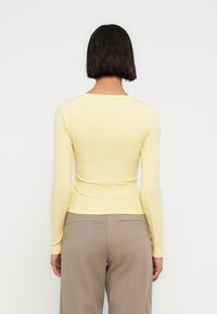 Ριμπ τοπ με μακριά μανίκια σε απαλό κίτρινο, εφαρμοστή γραμμή με στρογγυλή λαιμόκοψη, συνδυασμένο με ανοιχτό μπεζ παντελόνι.  Λείο ύφασμα, μίνιμαλ λεπτομέρειες.
