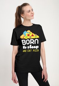 Joven mujer vestida con una camiseta negra que tiene un trozo de pizza de dibujos animados, un oso azul y el texto "Nacida para dormir y comer pizza", mirando hacia arriba y sonriendo.