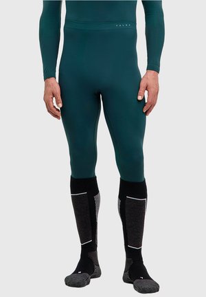 Uomo che indossa una maglia aderente a maniche lunghe verde scuro e leggings, con calze alte al ginocchio nere e grigie, in piedi contro uno sfondo semplice.