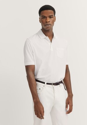 Weißes Polo-Shirt mit einer Knopfleiste mit zwei Knöpfen, kurzen Ärmeln und einer Brusttasche, kombiniert mit weißen Hosen und einem geflochtenen braunen Gürtel.
