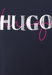 Tissu bleu marine avec le mot "HUGO" écrit en grandes lettres blanches majuscules avec une ligne rose en italique qui serpente à travers le texte.