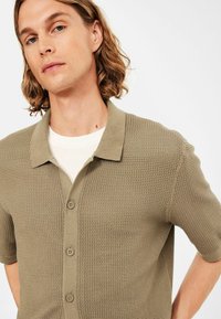 Man met schouderlang golvend haar, gekleed in een beige textuur kortarm overhemd met knopen over een wit T-shirt tegen een effen achtergrond.