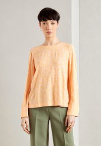 Haut à manches longues en tissu orange doux avec un petit motif floral blanc, présentant un col rond et une coupe décontractée, associé à un pantalon vert.
