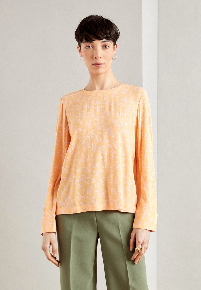 Haut à manches longues en tissu orange doux avec un petit motif floral blanc, présentant un col rond et une coupe décontractée, associé à un pantalon vert.