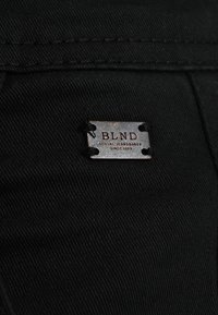 Gros plan sur un tissu en denim noir avec une petite étiquette métallique rectangulaire cousue, portant l'inscription "BLND Global Jeansmaker Depuis 1983".