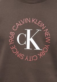 Hnedá mikina s okrúhlym logom obsahujúcim nápisy "CALVIN KLEIN NEW YORK" a "1968" v ružovej farbe a výrazným bielym "cK" v strede.