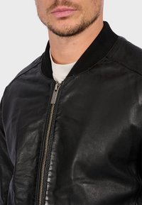 Černá kožená bomber bunda se zipem vpředu, žebrovaným límcem a hladkým povrchem. Má minimální švy a přiléhavý střih.