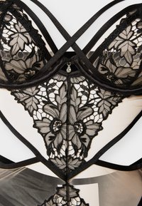 Μαύρο δαντελένιο εσώρουχο με λουλουδάτα μοτίβα, διάφανα τμήματα από δίχτυ, σταυρωτούς ιμάντες στους ώμους και ένα μικρό δάκρυ σε σχήμα κόσμημα στο κεντρικό μπροστινό μέρος.