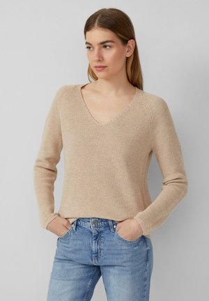 Beige rebrovaný pletený sveter s výstrihom do V, dlhými rukávmi a voľným strihom, v kombinácii so svetlomodrými džínsami.
