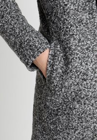 Abrigo de lana gris con un acabado texturizado y moteado. Presenta un bolsillo lateral y mangas entalladas con un sutil detalle de puño.