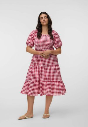 Femme debout, mains jointes, portant une robe à carreaux rouges et blancs avec des manches bouffantes et des sandales beiges sur un fond uni.