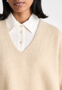 Beige knitted sweater with a V-neck, layered over a white, collared button-up shirt featuring subtle vertical stripes.