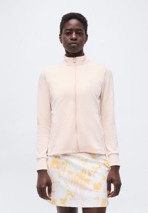 Άτομο που φοράει ένα ανοιχτό ροζ ζακετάκι με φερμουάρ και ριμπ ύφασμα και μια λευκή φούστα με κίτρινα-πορτοκαλί αφηρημένα λουλουδένια σχέδια, στέκεται μπροστά σε ένα απλό φόντο.