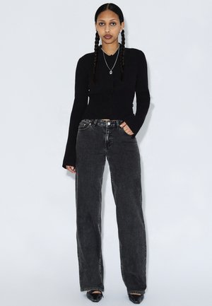 Schwarzer Rippstrick-Cardigan, kombiniert mit locker sitzenden schwarzen Denim-Jeans mit hoher Taille und leicht ausgefranstem Saum. Komplettiert mit schwarzen Schuhen.