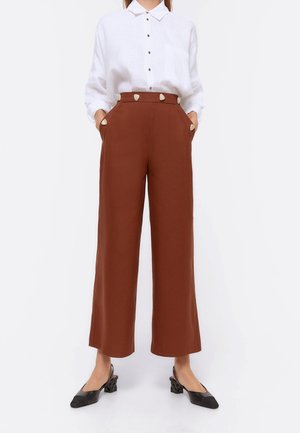 Mujer con pantalones marrones de talle alto y piernas anchas, adornados con botones decorativos, y camisa blanca abotonada, combinados con zapatos negros de tacón.