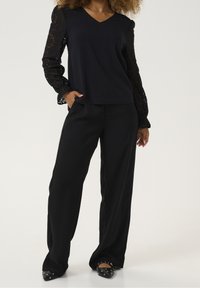 Czarna bluzka z jedwabnym tułowiem i koronkowymi rękawami, z dekoltem w serek, zestawiona z czarnymi spodniami o szerokich nogawkach oraz płaskimi butami w szpic z ćwiekami.
