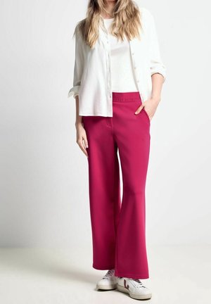 Kvinde iført hvid skjorte med knapper, hvid top, magenta vidde bukser og hvide sneakers, stående med den ene hånd i lommen.