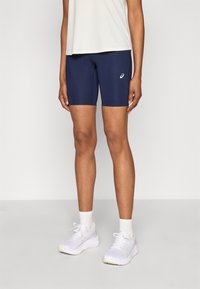 Σκούρο μπλε αθλητικό σορτς με λείο ύφασμα και εφαρμοστή σχεδίαση, που φέρει λευκό λογότυπο στο μηρό, σε συνδυασμό με λευκά αθλητικά παπούτσια.