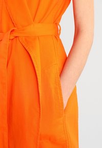 Orange beklædning med en taljebånd, der har en lomme detalje. Stoffet virker glat og har en let tekstur.