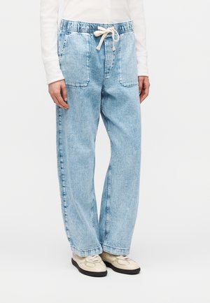 Άτομο που φορά ανοιχτό μπλε φαρδιά τζιν παντελόνια με ελαστική μέση και κορδόνι, λευκό μακρυμάνικο τοπ και κρεμ παπούτσια με μαύρες σόλες.