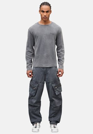 Giovane uomo con capelli intrecciati che indossa una maglietta grigia a maniche lunghe, pantaloni cargo larghi e sneakers bianche, in piedi davanti a uno sfondo semplice.