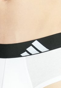 Weiße Unterwäsche mit einem schwarzen elastischen Bund, der drei weiße Streifen und ein adidas-Logo auf der linken Seite zeigt. Glatte Textur.
