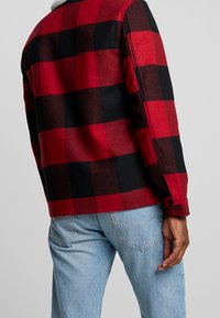 Henkilö, jolla on punamusta ruudullinen takki ja vaaleansiniset farkut, kuvattuna takaa yksiväristä taustaa vasten.