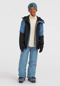 Ski outfit featuring a blue and black jacket, cream fleece layer, blue pants, black gloves, and blue beanie, with gray ski boots.