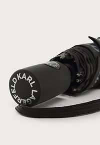 Parapluie noir avec un manche cylindrique à finition mate, un logo imprimé en blanc et une sangle texturée pour une meilleure prise.