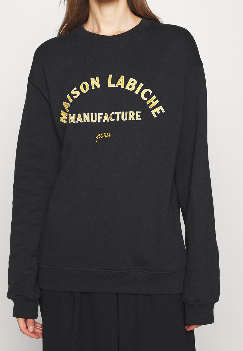 Personne portant un sweat-shirt noir avec un texte jaune « MAISON LABICHE MANUFACTURE paris » centré sur la poitrine.