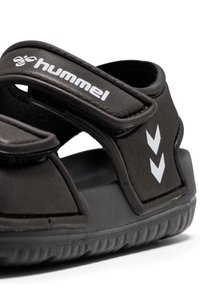 Sandale noire avec straps Velcro, semelle en caoutchouc et dessus texturé. Présente un logo blanc et des accents en chevron sur le côté.